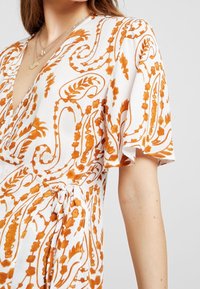 Robe blanche avec un motif floral et paisley orange, présente un décolleté en V, de courtes manches à volant et un détail de taille nouée.