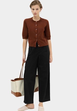 Ung kvinde iført en brun kortærmet striktrøje med knapper, brede sorte bukser, beige slip-on sko og bærende på en stor beige og brun tote-taske.