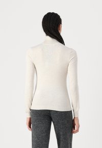 Pulover bej cu guler înalt, design ajustat, manșete și tiv în relief, textură netedă, material ușor, purtat cu pantaloni gri.