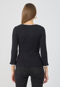Svart langermet topp med bølget hals og mansjetter, laget av ribbestrikket stoff. Designet har en tettsittende fasong, kombinert med mørke jeans.