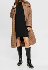 Cappotto lungo trapuntato marrone sopra un abito nero lungo fino al ginocchio, abbinato a stivali neri lucidi al ginocchio con suole spesse e resistenti.