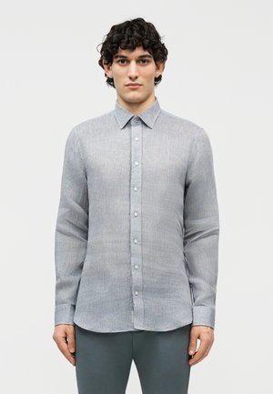 Άνδρας με σγουρά σκουρόχρωμα μαλλιά φορώντας εφαρμοστό ανοιχτό γκρι πουκάμισο με κουμπιά και σκούρα γκρι παντελόνι, στέκεται μπροστά σε απλό λευκό φόντο.