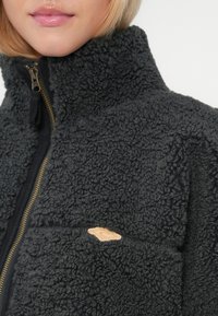 Schwarze Fleecejacke mit strukturiertem Material, ausgestattet mit einem hohen Kragen, einem Reißverschluss auf der Vorderseite und einem kleinen tanfarbenen Logo-Akzent auf der Brusttasche.
