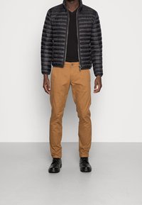 Chaqueta de plumas negra con un acabado brillante, combinada con pantalones chinos color marrón. Zapatos negros de cordones. Camiseta negra de cuello en V simple por debajo.