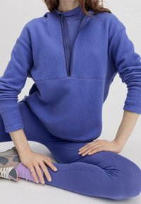 Lila fleecepullover med halvdragkedja fram, bärs över matchande leggings. Mjuk textur, avslappnad passform, ribbade muddar och lager-på-lager högkrage design.