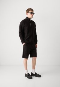 Sweatshirt zippé noir, shorts noirs, chaussures noires à enfiler, chaussettes blanches et grandes lunettes de soleil noires. Design minimaliste et palette monochromatique.