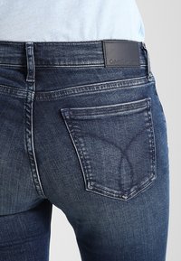 Tummansiniset farkut, joissa on räätälöity istuvuus, hienovarainen kuluminen ja nahkainen Calvin Klein -logo vyötäröllä. Erityinen takataskun muotoilu.