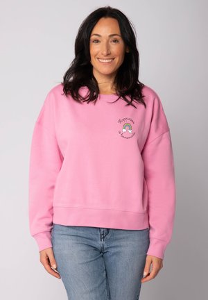 Sweatshirt rose à col ras du cou avec des poignets et un ourlet côtelés. Présente le texte "Le bonheur se fait maison" et une broderie arc-en-ciel sur la poitrine.