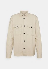 Beżowa koszula zapinana na guziki, wykonana z miękkiego materiału. Posiada dwie kieszenie na piersi, czarne guziki i klasyczny kołnierz. Długie rękawy z zapięciami na guziki.