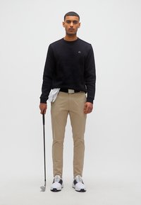 Άνδρας με μαύρο πουλόβερ και μπεζ παντελόνι κρατάει μπαστούνι γκολφ με άσπρα γάντια κρυμμένα στη ζώνη του, στέκεται μπροστά σε λευκό φόντο.