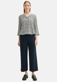 Schwarz-weiß gemusterte Bluse mit V-Ausschnitt, kombiniert mit weiten marineblauen Hosen und klobigen schwarzen Schuhen.