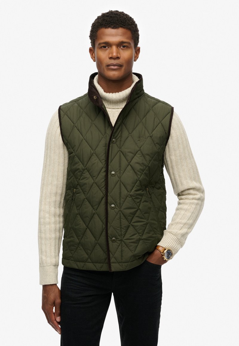 Gilet matelassé vert avec col montant, bordure marron et boutons-pression. Porté par-dessus un pull en laine crème, avec des poches latérales.
