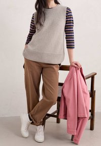 Helles graues, gestricktes ärmelloses Oberteil über einem gestreiften Langarmshirt, kombiniert mit braunen Hosen und weißen Turnschuhen. Eine rosa Jacke liegt über einem Stuhl.