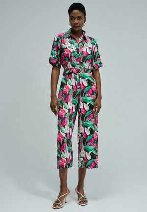 Femme portant une combinaison à manches courtes ceinturée avec un motif floral rose, blanc et vert, associée à des talons blancs à brides, debout devant un fond uni.
