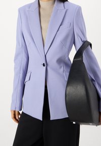 Persona che indossa un blazer viola chiaro sopra un top beige a coste, pantaloni neri e porta una grande borsa in pelle nera sul braccio sinistro.