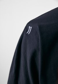 Black cotton shirt featuring a smooth texture. A small, light gray logo is positioned on the shoulder. Seam detail visible.