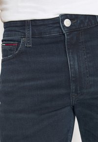 Tummanpesu denim-farkut, joissa on normaali istuvuus. Niissä on edessä nappisuljenta, vetoketju ja punaiset yksityiskohdat etutaskussa.