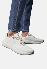 Sneaker atletici bianchi con un design elegante, materiale strutturato e dettagli crema. Presentano lacci beige e una suola grigia.