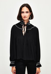 Schwarze Bluse mit hohem, rüschengeschmücktem Kragen, verziert mit weißer Stickerei und einem Bindedetail. Lange Ärmel mit gemusterten Manschetten. Weicher Stoff, lockere Passform.