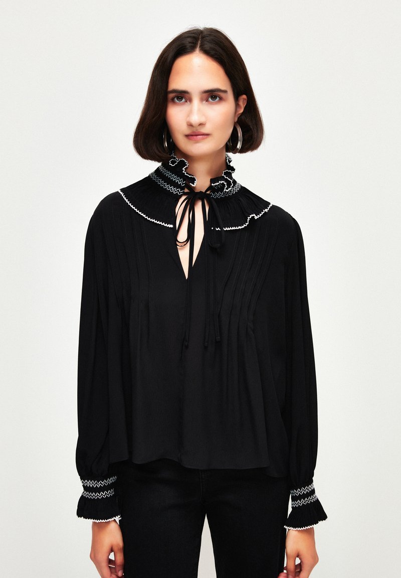 Schwarze Bluse mit hohem, rüschengeschmücktem Kragen, verziert mit weißer Stickerei und einem Bindedetail. Lange Ärmel mit gemusterten Manschetten. Weicher Stoff, lockere Passform.