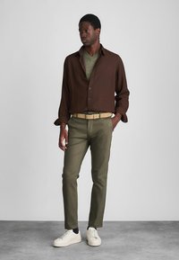 Camicia marrone a bottoni sopra una maglietta verde a scollo a V, abbinata a pantaloni verde oliva e sneaker crema. Presenta una cintura beige e uno sfondo neutro.