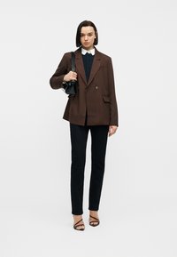 Γυναίκα με καφέ σακάκι πάνω από μαύρο τοπ και παντελόνι, κρατώντας μαύρη τσάντα ώμου, στέκεται μπροστά σε λευκό απλό φόντο.