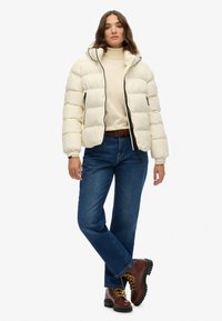 Pufferjack in off-white, met een ritssluiting aan de voorkant, capuchon en zwarte accenten. Gecombineerd met blauwe jeans en bruine veterschoenen.
