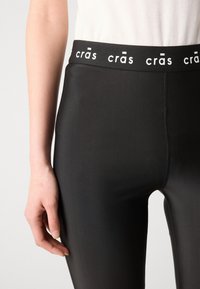 Crne tajice s glatkom teksturom i prilagođenim dizajnom, s pojasom ukrašenim logom u kontrastnoj bijeloj boji.