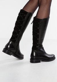 Botas negras hasta la rodilla con un frente de cuero liso y parte trasera de ante, que cuentan con cordones entrecruzados, una cremallera lateral y un tacón bajo.