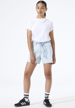Vrouw staand, gekleed in een wit t-shirt, lichtblauwe denimshorts met een gestrikte riem, witte sokken met zwarte strepen en zwarte sneakers.