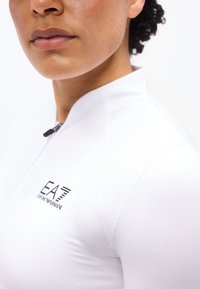 Γυναίκα που φοράει λευκή μπλούζα με φερμουάρ και μαύρο λογότυπο EA7 Emporio Armani στο στήθος, κοντινή λήψη στο πρόσωπο και το πάνω μέρος του κορμού.