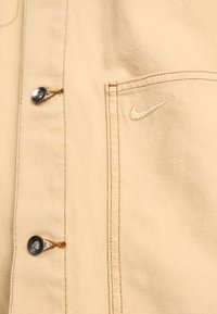 Beige bomullsjacka med en slät textur, som har en knappstängning och en guldbroderad Nike-logga på bröstfickan.
