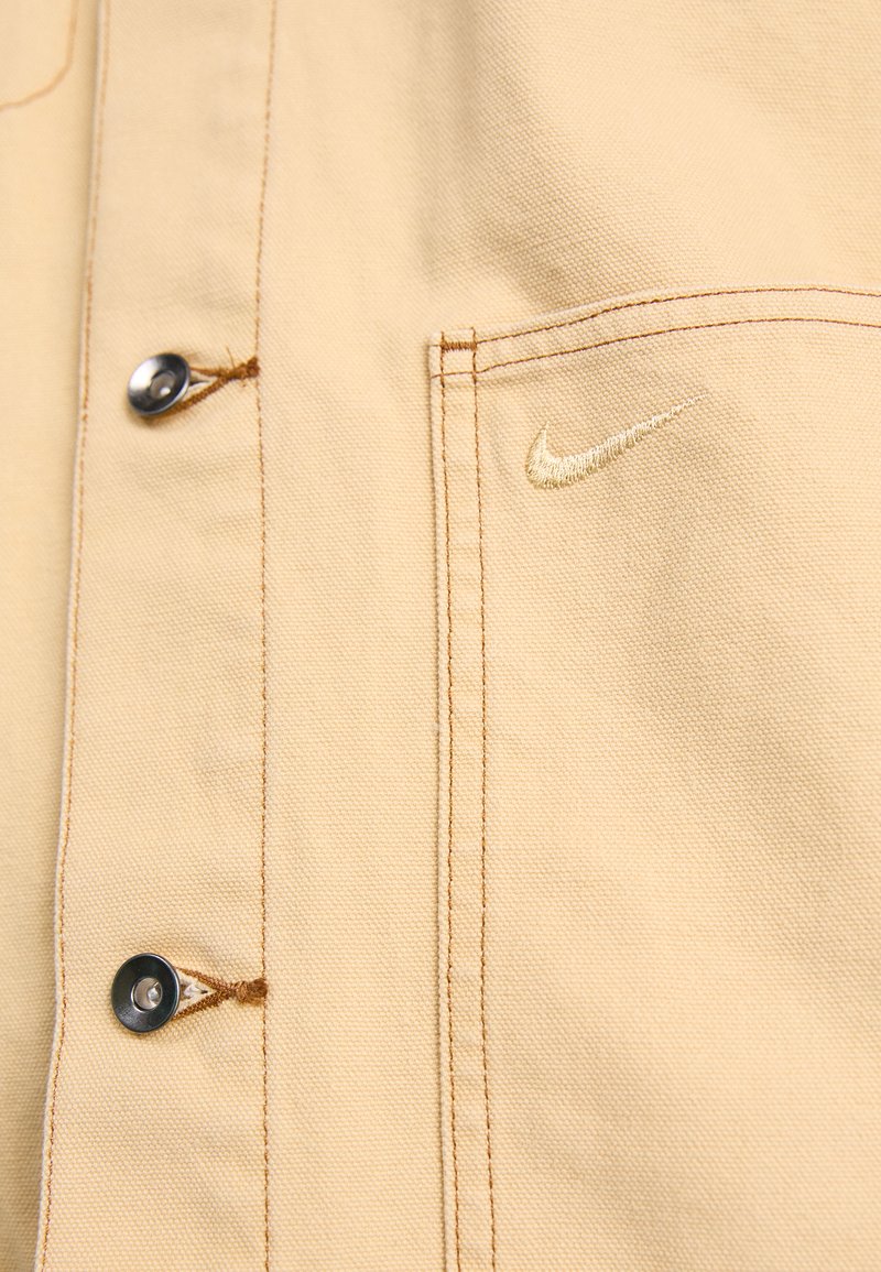Chaqueta de algodón beige con una textura suave, destaca por su cierre de botones y un logo de Nike bordado en dorado en el bolsillo del pecho.