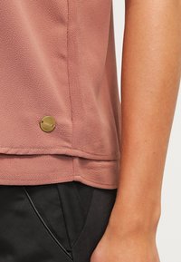 Haut à manches courtes rose poudré en tissu texturé avec un ourlet arrondi ; présente un accent logo circulaire doré sur le bas à gauche.