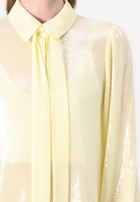 gele paillettenblouse met een puntige kraag en strikdetail; heeft een doorschijnende textuur en een glanzend, reflecterend oppervlak.