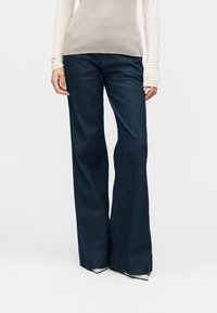 Jeans di denim blu scuro a gamba larga con una texture liscia, caratterizzati da vita alta e orlo risvoltato. Abbinati a una maglia leggera a maniche lunghe.