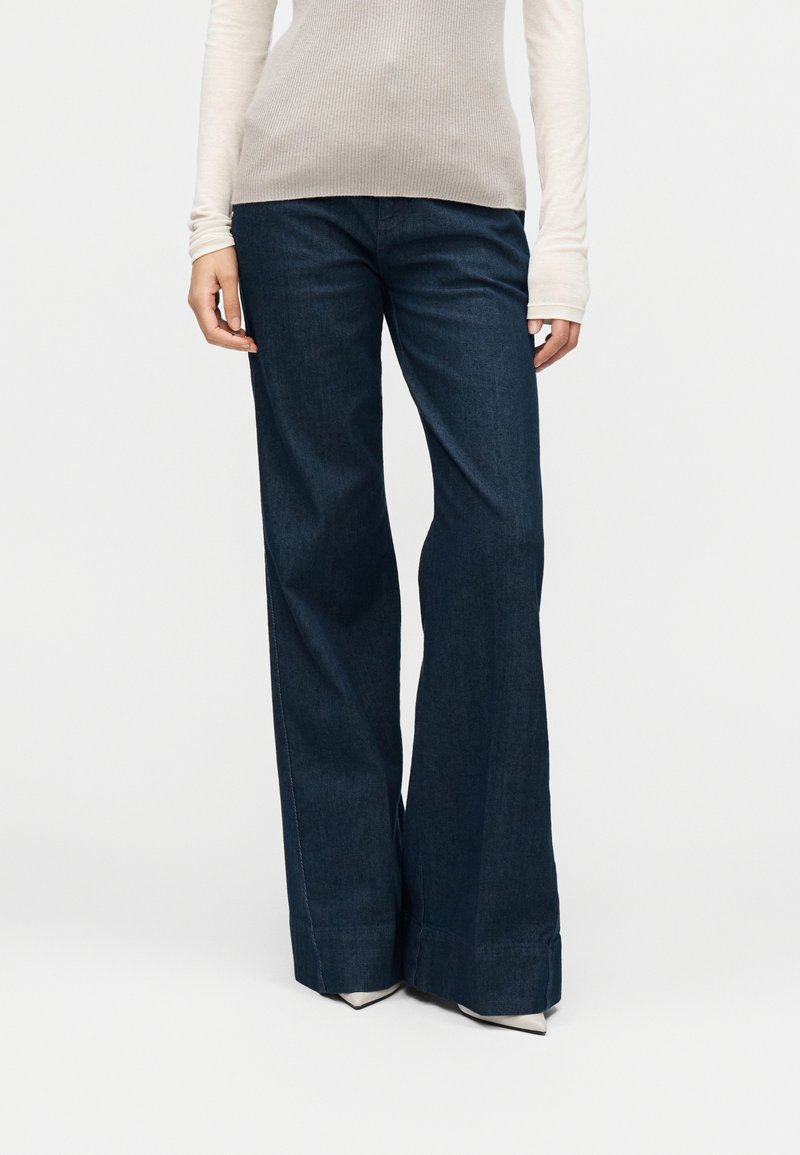 Jeans di denim blu scuro a gamba larga con una texture liscia, caratterizzati da vita alta e orlo risvoltato. Abbinati a una maglia leggera a maniche lunghe.