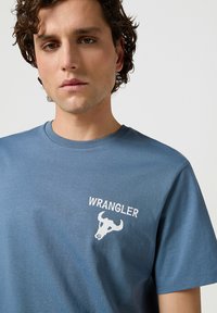 Camiseta de algodón azul con mangas cortas que presenta un logo blanco de "WRANGLER" y un gráfico de un cráneo de toro en la parte izquierda del pecho.