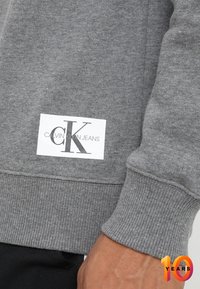 Felpa grigia con polsini e orlo a costine, dotata di un'etichetta bianca che mostra il logo "CALVIN KLEIN JEANS" in nero.