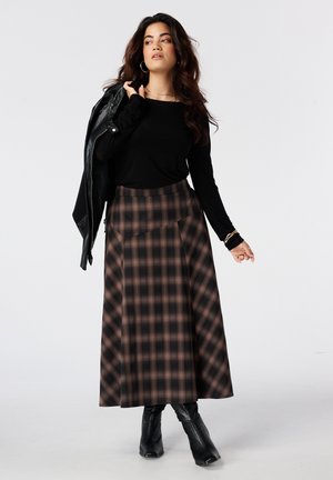 Vrouw draagt een zwarte top met lange mouwen, een lange bruine geruite rok, zwarte laarzen en houdt een zwarte leren jas over haar schouder.