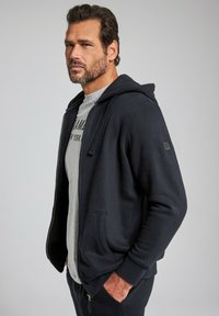 Marinefarbener Kapuzenpullover mit Reißverschluss, einer Fronttasche, einer Kapuze mit Zugband und einem grauen T-Shirt darunter mit aufgedrucktem Logo. Aus weichem Material, entspannter Schnitt.