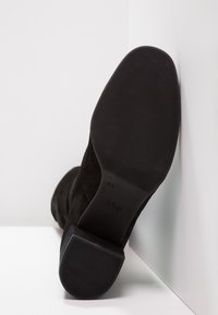 Bota de tobillo de ante negro con una suela de goma texturizada, que presenta un diseño elegante y un tacón curvado para mayor estabilidad. Logotipo de la marca visible en la suela.