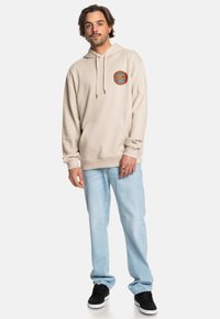 Béžová mikina s kapucňou vyrobená z bavlnenej zmesi, s prednou vreckom a farebným kruhovým logom na hrudi. V kombinácii so svetlomodrými džínsami.