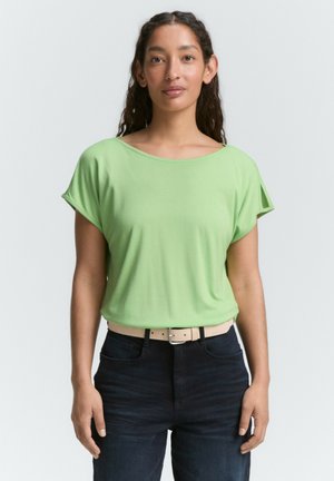 Top verde a maniche corte in tessuto morbido con scollatura ampia, abbinato a jeans blu scuro e una cintura di colore chiaro. Design semplice, texture liscia.