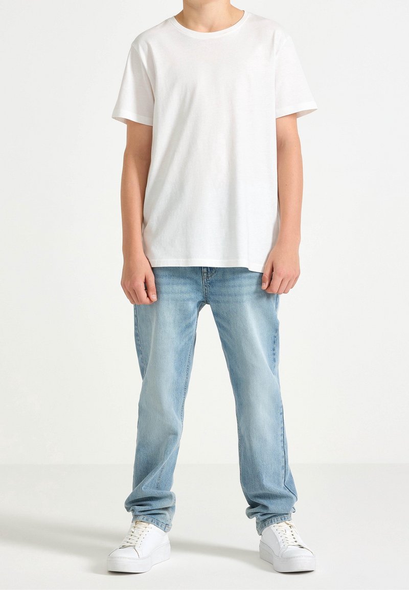 Personne portant un t-shirt blanc à manches courtes, un jean bleu clair et des baskets blanches, debout devant un fond blanc uni.