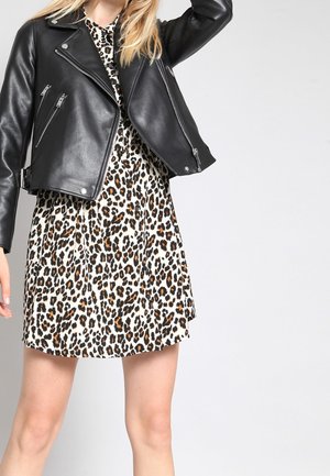 Blonde person wearing a black leather jacket over a leopard print dress, standing against a plain light gray background.