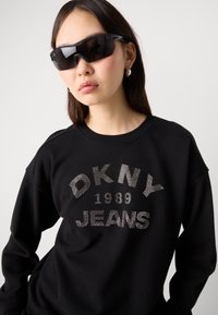 Svart hettegenser med "DKNY Jeans 1989" i metalliske nagler. Modellen har på seg sporty svarte solbriller og sølvfargede hoop-øreringer. Jevnt tekstur.
