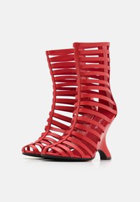 Punaiset remmillä varustetut korkokengät, joissa on ainutlaatuinen leikkausdesign. Valmistettu sileästä materiaalista, jossa on kaareva korko ja avoin varvas. Musta pohja.