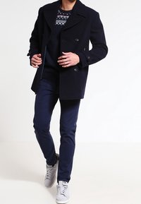 Cappotto doppiopetto blu navy con ampi risvolti, indossato sopra un maglione scuro con motivo bianco e blu, abbinato a pantaloni navy e sneakers grigie chiare.