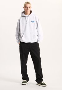 Felpa grigia con logo blu, tasca a marsupio e cappuccio con coulisse, abbinata a pantaloni neri e sneaker verdi con lacci bianchi.